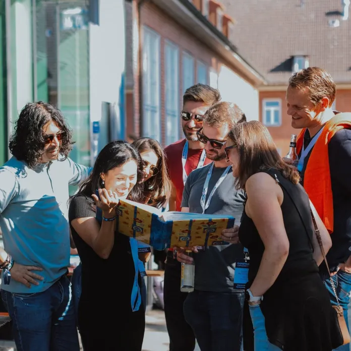 A group of attendees smile while looking through a colorful booklet together outside a campus building