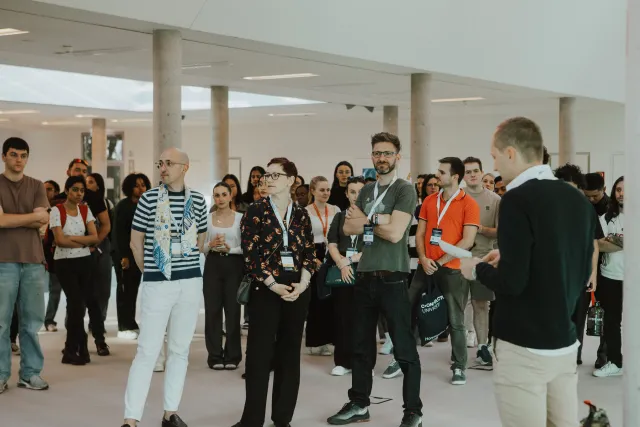 Attendees stand together and listen to a speaker during an indoor university event
