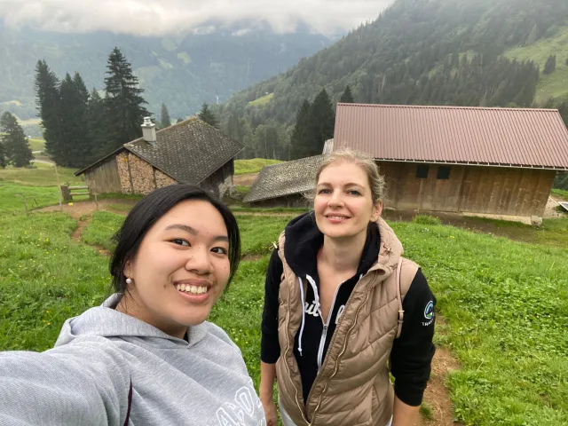 Two women pose for a selfie in a grassy mountain valley with wooden cabins and forest behind them